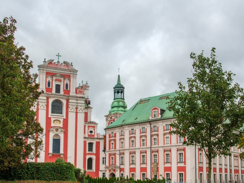 Na zdjęciu znajduje się barokowy kompleks budynków o różowo-białej elewacji. Po lewej stronie widoczna jest fasada kościoła z dużymi oknami, białymi zdobieniami i zielonym krzyżem na szczycie. Po prawej stronie znajduje się przylegający budynek klasztorny lub pałacowy z zielonym dachem i licznymi oknami. Na dachu wyróżnia się wieżyczka zwieńczona kopułą i krzyżem. Na pierwszym planie rosną drzewa i krzewy, a w tle widać pochmurne niebo.